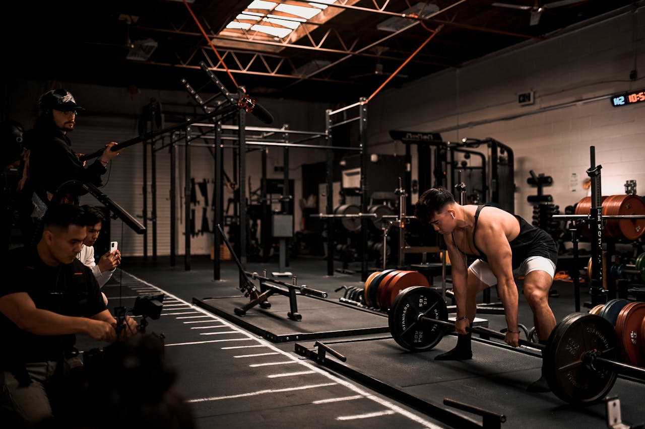 Muscular man performs deadlift in a gym with film crew documenting, showcasing strength and focus.